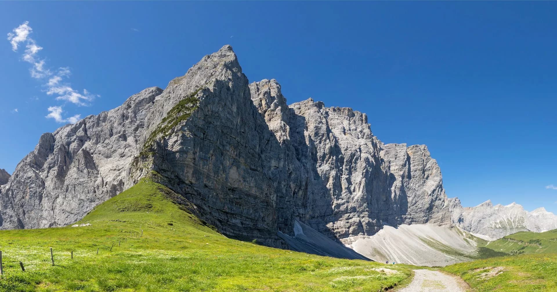 Rocky mountain peaks tower over a grassy alpine meadow with a dirt path leading toward the base.