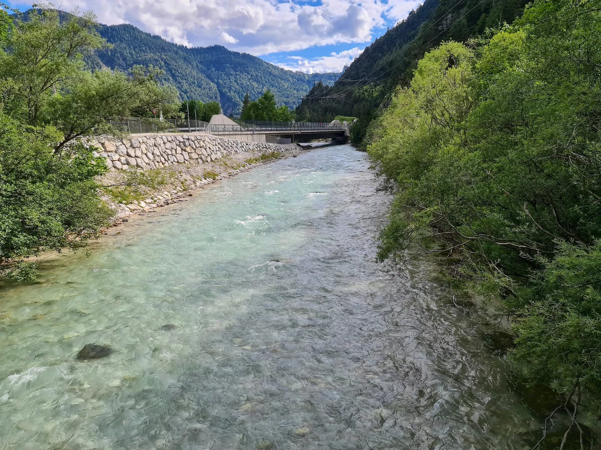 Clear river flowing through a valley near Scharnitz with forested mountains and a bridge.