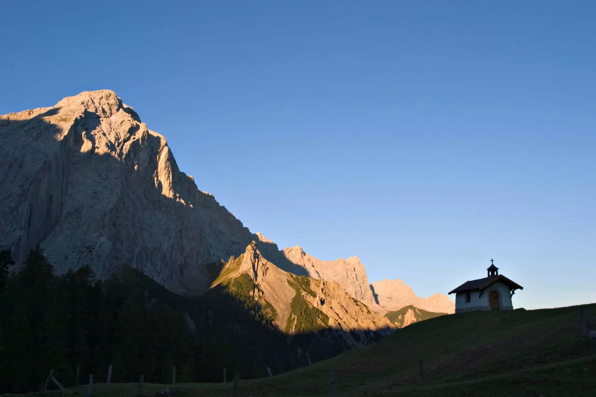 Small chapel on grassy hill below sunlit alpine mountains under clear blue sky.