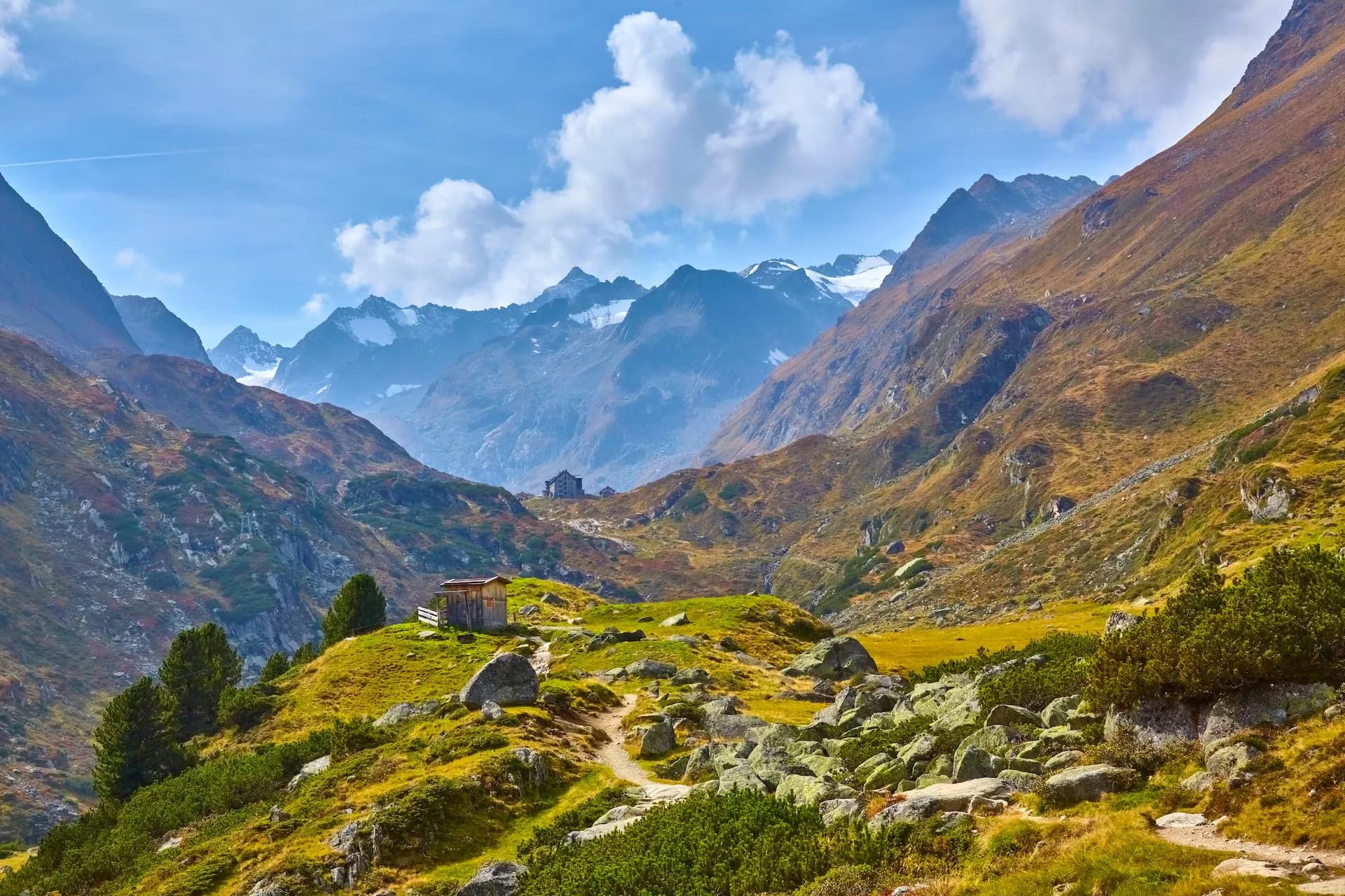 Alpine valley with hiking path, wooden hut, and snow-capped mountains under blue sky in Stubai Valley.