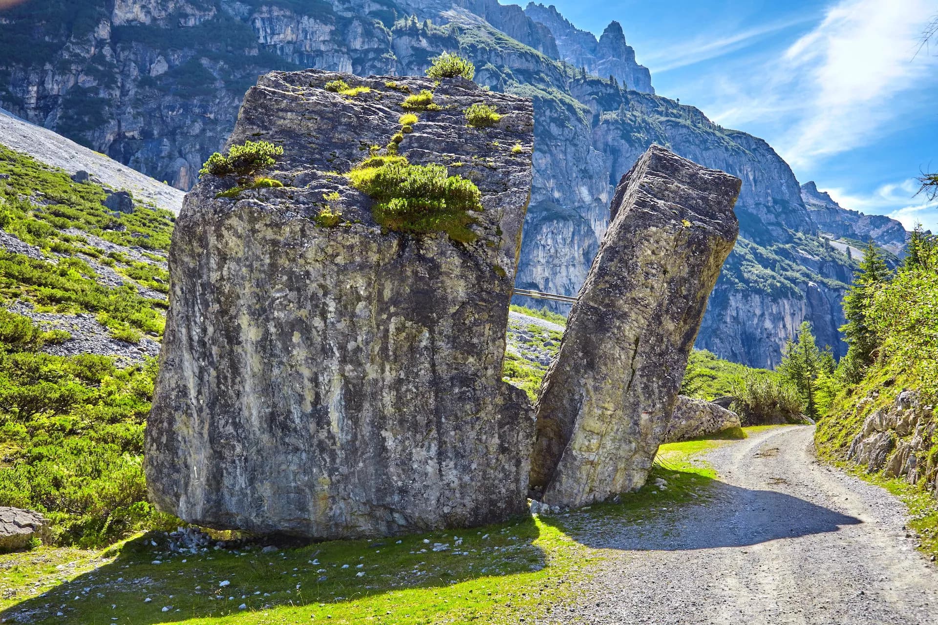 Split glacial erratic rock beside gravel path with steep, green mountains under blue sky.